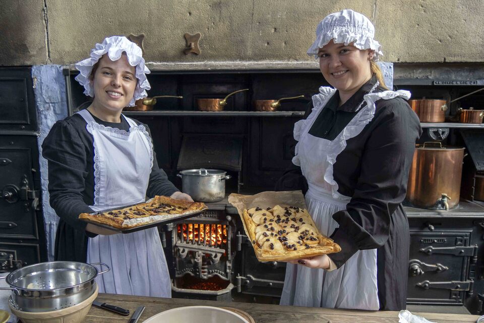 Two individuals in historical cook attire presenting trays of fruit-topped pastries in front of an old-fashioned kitchen stove, surrounded by copper pots and traditional kitchenware.